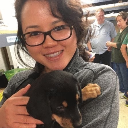 A woman wearing glasses is smiling while holding a black and tan puppy in her arms.
