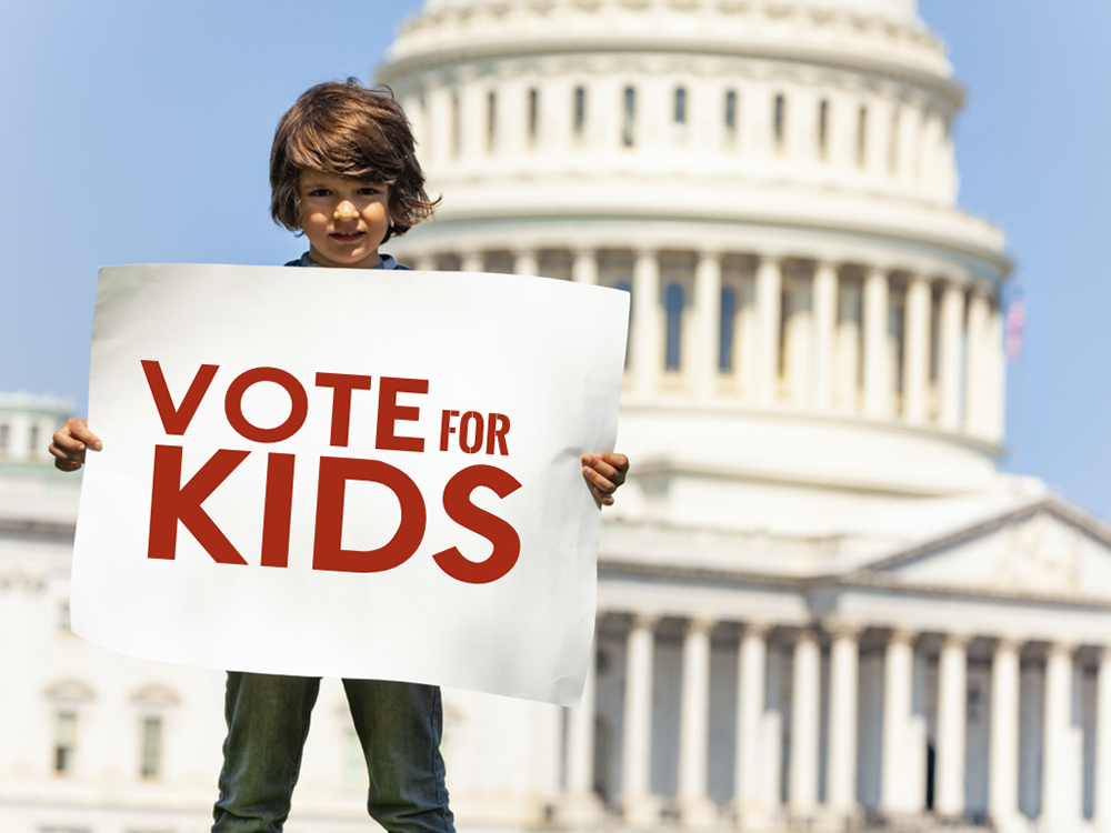 A child holds a sign that reads "Vote for Kids" in front of a government building