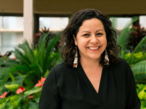 This image shows Carly Bad Heart Bull's headshot. She stands outside in front of greenery, smiling.