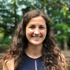 Elizabeth Allaby is smiling in front of trees, and her brown hair is covering her shoulders.