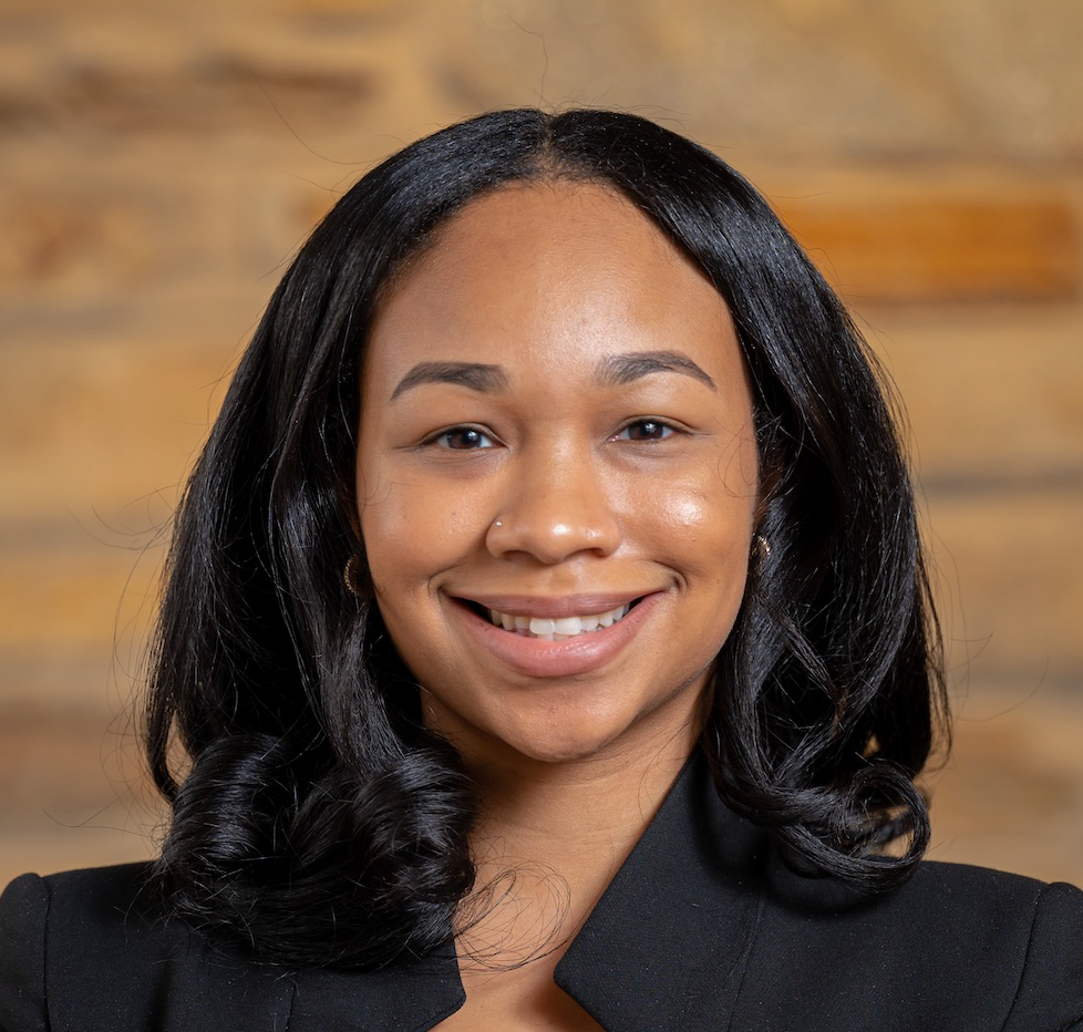 Rahmirah is wearing a black suit jacket and standing in front of a brick wall