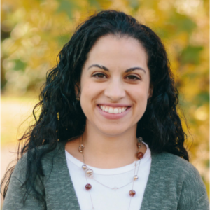 Grace Asenjo is standing outside, wearing a grey cardigan and a red necklace for her headshot photo