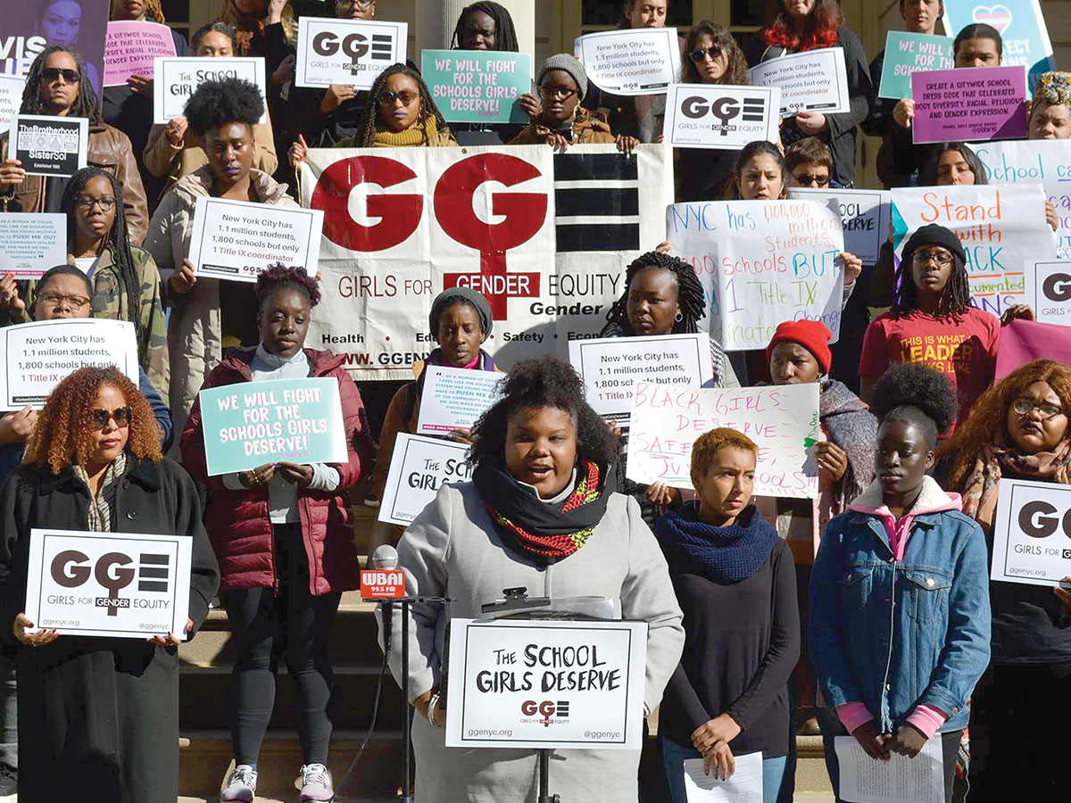 A young woman dressed in a gray jacket and colorful scarf addresses a crowd during a rally. Behind her stand a group of people holding "Girls for Gender Equity" signs.