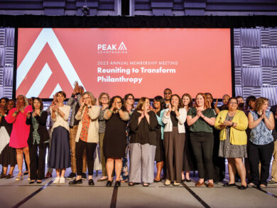 PEAK's staff and board of directors stand on stage facing the crowd and clap. Behind them is a red screen that reads "2023 Annual Membership Meeting: Reuniting to Transform Philanthropy."