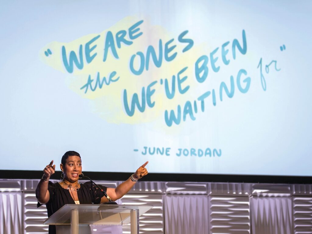 Satonya Fair points to the crowd during her welcome address at PEAK2023 in Baltimore. Behiind her is a screen that reads "We are the ones we've been waiting for" in blue handwriting.