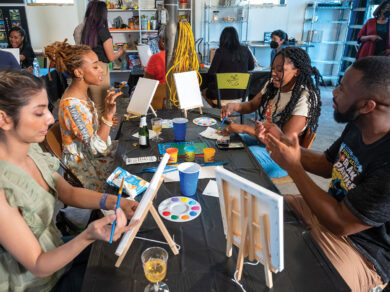 A group of people talk and paint over a table full of painting supplies.