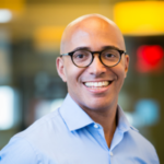 Jason is smiling in his headshot. He is wearing black glasses and a light blue collared shirt