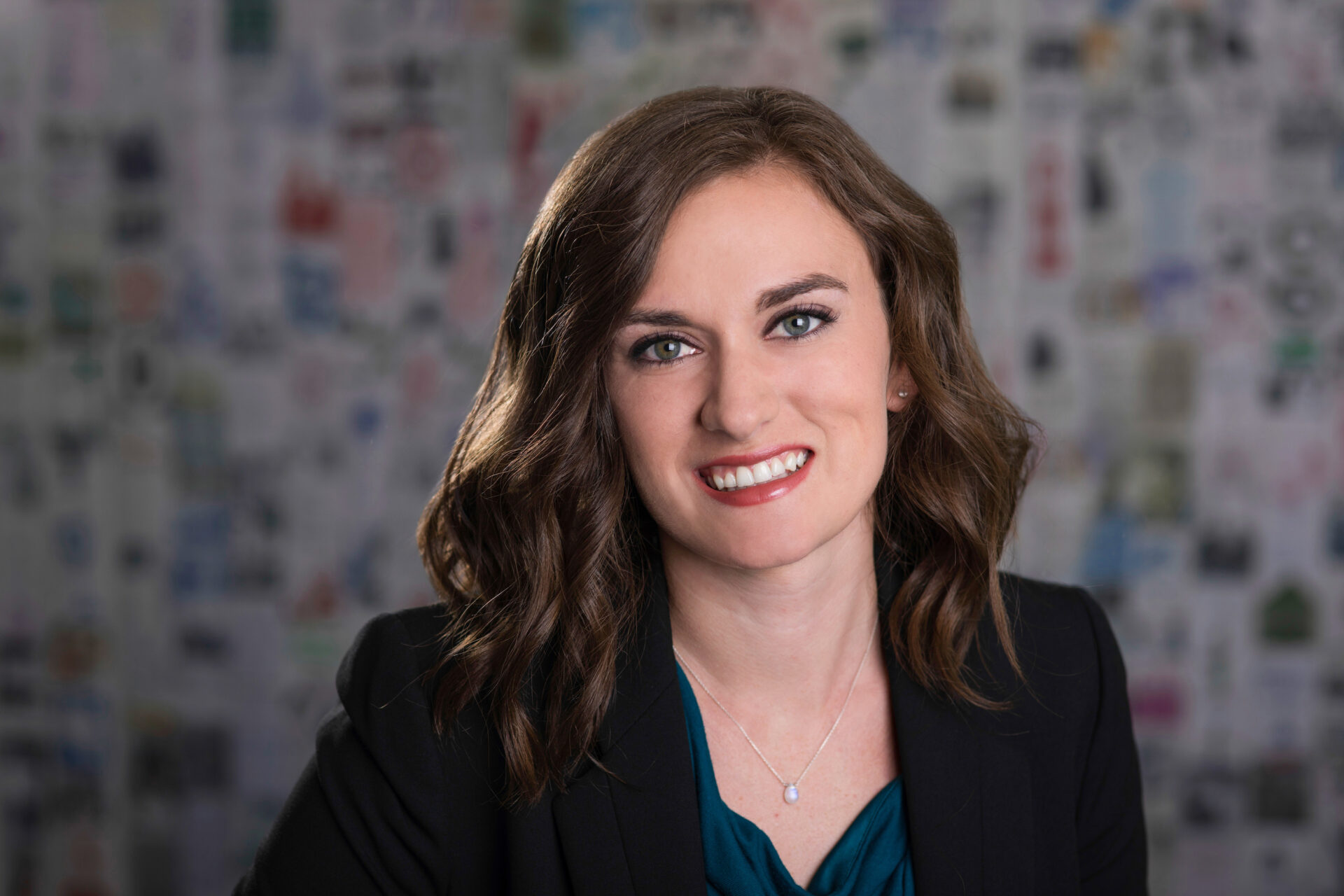 Kelli Rojas smiles for her headshot picture in front of a multicolored background, wearing a black blazer and blue blouse.