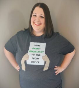 Kristen Summers is a light skinned woman wearing bright red lipstick and a shirt that reads "bring grants management to the front office" in black and green text.