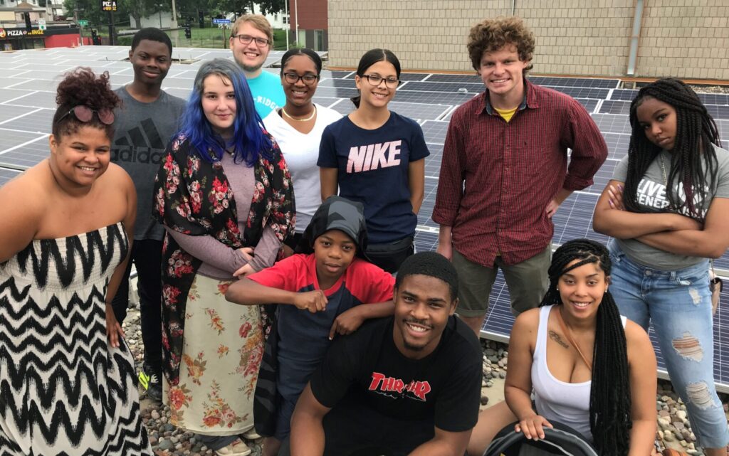 A group of kids smiles for a photo in front of solar panels