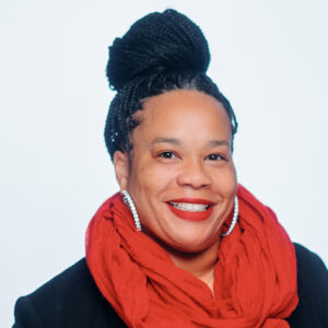 Miyesha Perry smiling in her photo, wearing a black jackets and bright red scarf with silver hoop earrings