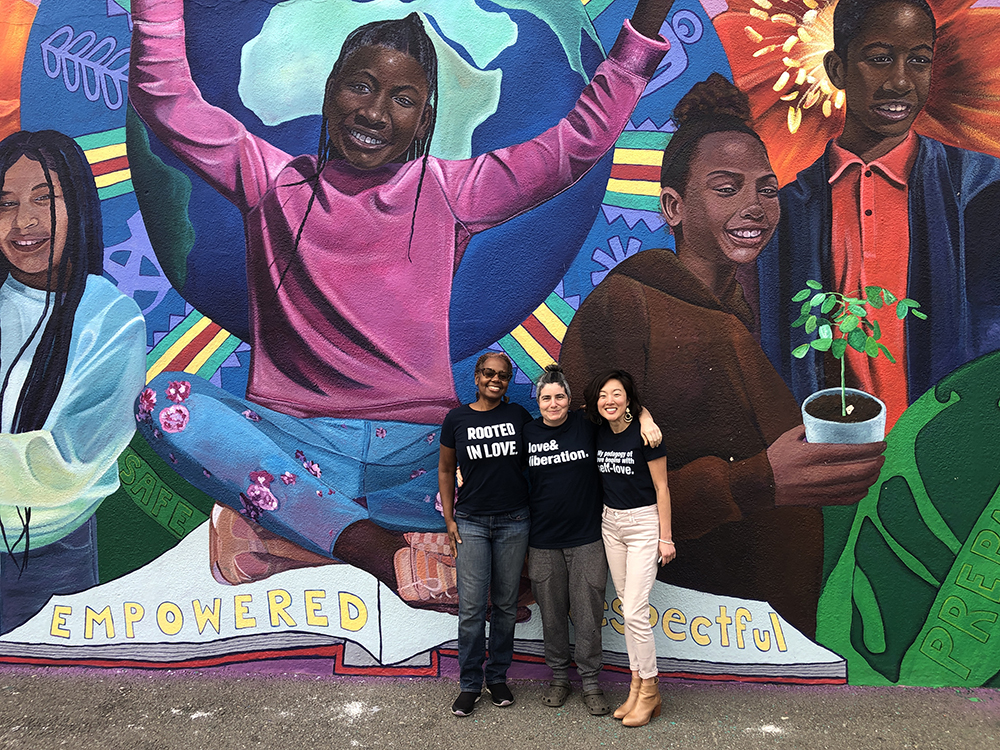 The Agency by Design Oakland team outdoors, standing in front of a colorful mural featuring happy, young Black children.