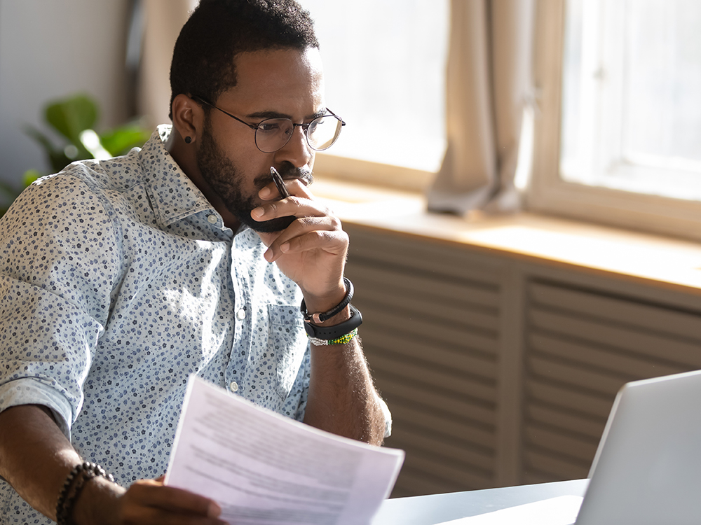 A Black male professional sits at his desk studying documents and information on his computer screen
