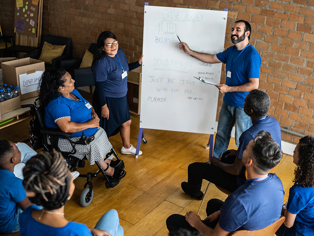 An adult man and woman in front of a large whiteboard leading a conversation with seven colleagues, one of whom is a wheelchair user