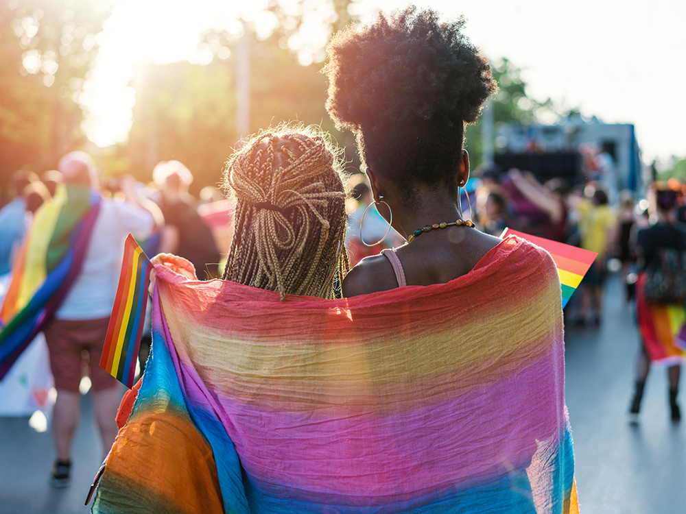A young couple stand with their backs to the camera with a colorful piece of fabric draped over their shoulders, while holding a small rainbow flag.