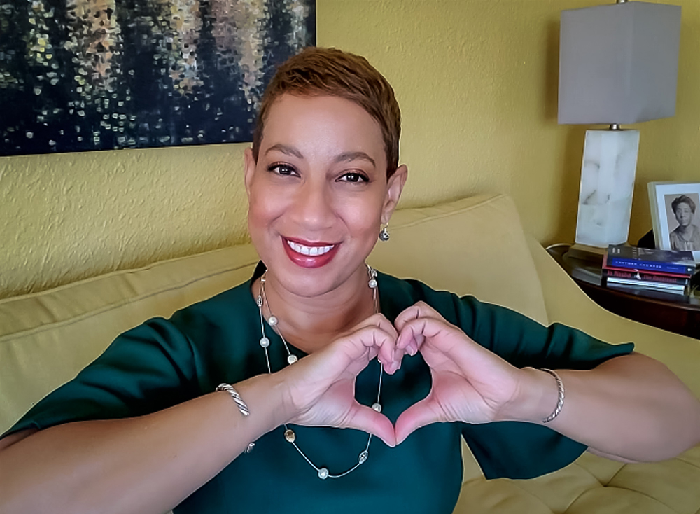 Satonya Fair is smiling, sitting on a yellow couch, and making a heart with her hands.