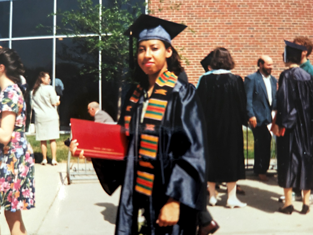 Satonya Fair is wearing a black graduation robe and cap with a kente cloth stole. She is holding a red diploma cover.