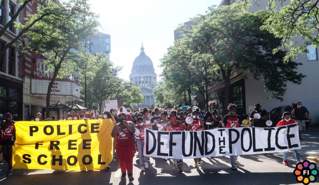 A crowd walks down a city street holding signs that read "police free school" and "defund the police."