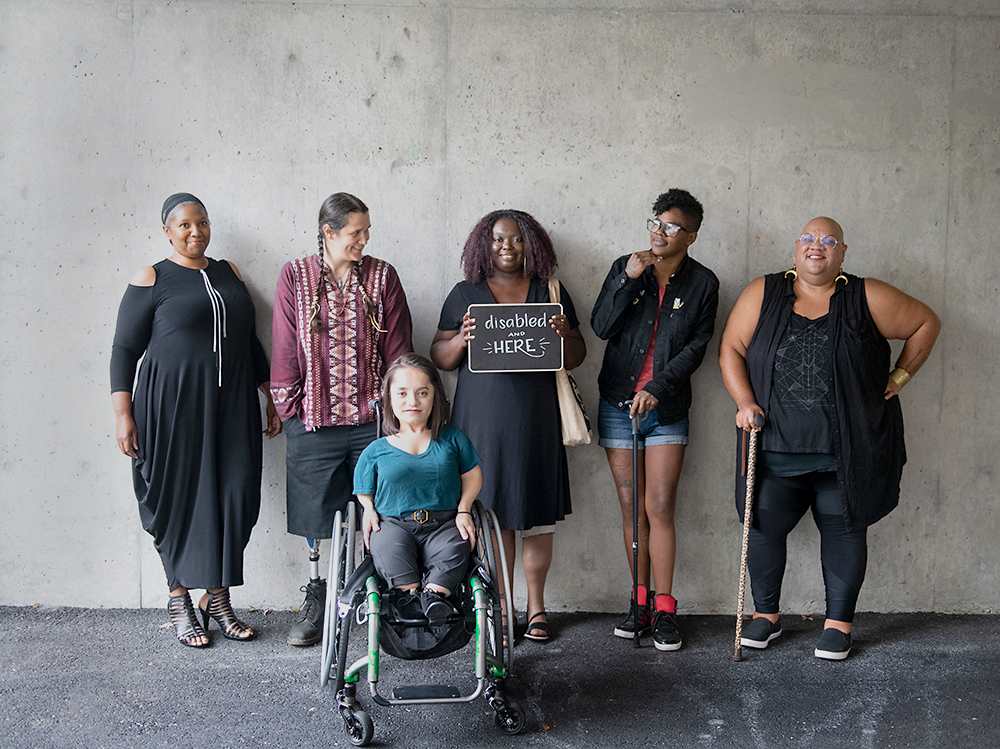 Six disabled people of color smile and pose in front of a concrete wall. Five people stand in the back, with the Black woman in the center holding up a chalkboard sign reading "disabled and here." A South Asian person in a wheelchair sits in front.
