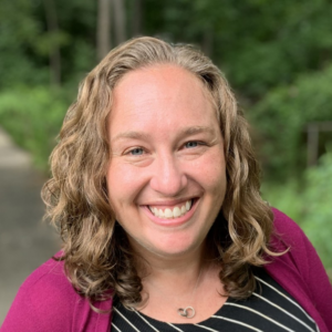 Marissa is smiling in her headshot. She is standing in front of green foliage. She is wearing a black top with white diagonal stripes and a dark fuchsia sweater