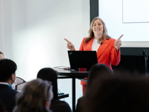 Jennifer Marsack wears a bright orange-red blazer and stands in front of a room while gesturing with her hands