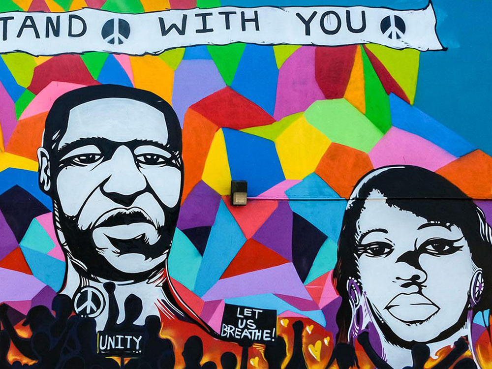 A mural shows victims of police brutality on a multicolor background. A crowd holes signs that read "Let us breathe" and "unity."