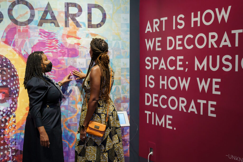 Two PEAK2023 attendees stand in front of an installation at Reginald F. Lewis Museum of African American History and Culture. Next to them, white text on a burgundy wall reads, "Art is how we decorate space; music is how we decorate time."