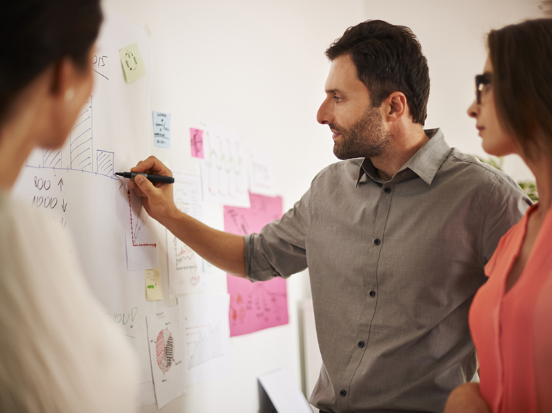 An executive writes out data on a dry erase board as two colleagues look on